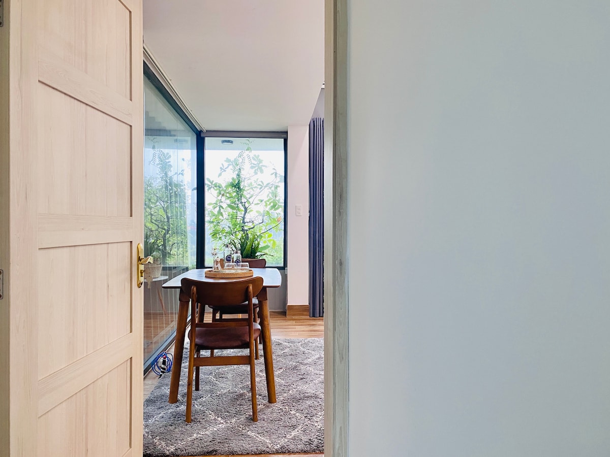 A doorway leads into a bright space featuring a round wooden dining table surrounded by three chairs. Large glass windows allow natural light to flood in, showcasing greenery outside. A soft rug covers the floor, adding comfort to the room's inviting atmosphere.