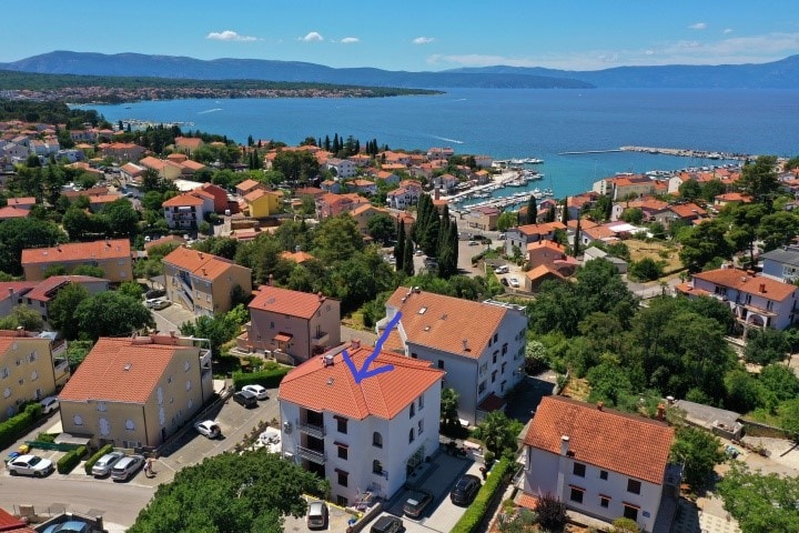 An aerial view of a coastal town illustrates a blend of residential buildings with red-tiled roofs. The blue waters of the nearby sea are visible, along with a small marina in the distance, surrounded by greenery and trees.