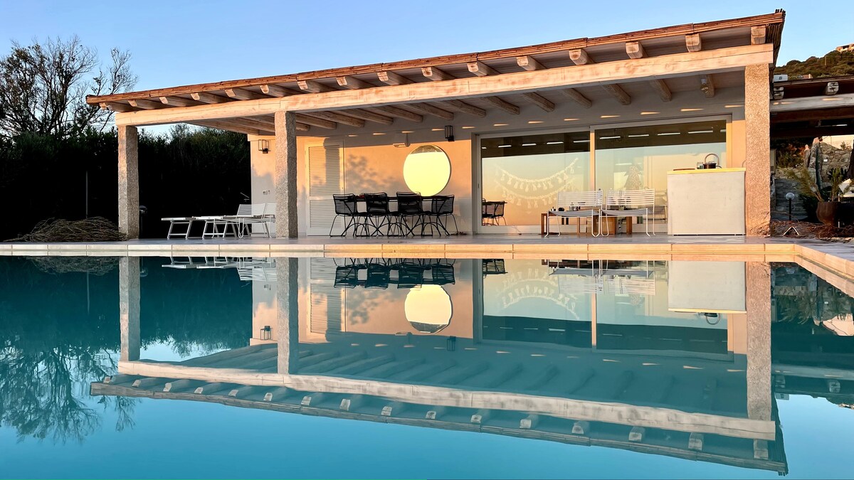 An elegant villa features an expansive outdoor area with a covered terrace and large dining table. The sleek infinity pool reflects the structure and surrounding nature, creating a serene atmosphere. Soft light highlights the clean lines and modern design, inviting relaxation and enjoyment.