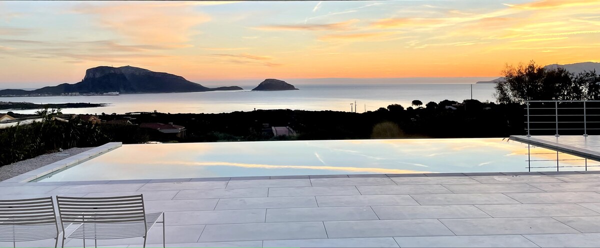 An infinity pool extends towards the horizon, reflecting the soft colors of a sunset. The ocean view is complemented by distant islands and a serene landscape. Two white chairs sit nearby on a tiled patio, providing a relaxing spot to enjoy the scenery.