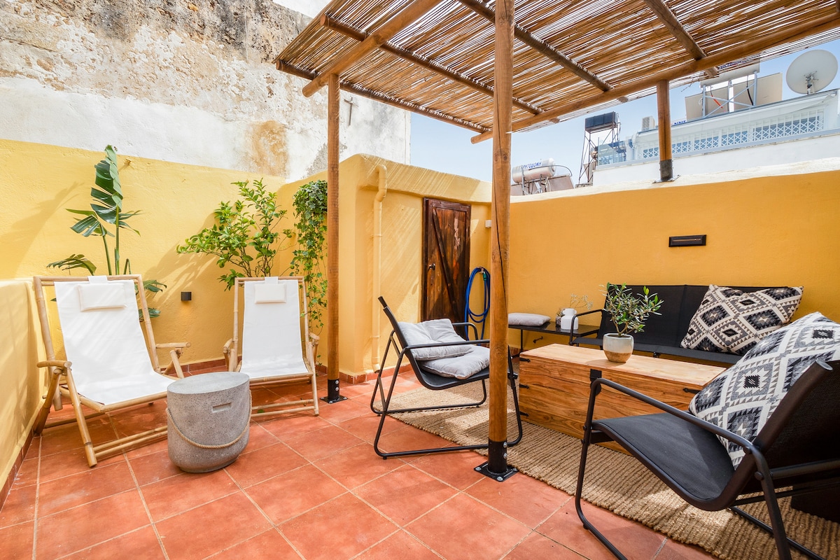 A private rooftop terrace features a mixture of seating options, including two lounges and a black armchair. A wooden coffee table is surrounded by greenery, with textured rugs covering the terracotta tiles. Yellow walls create a warm and inviting outdoor space for relaxation.