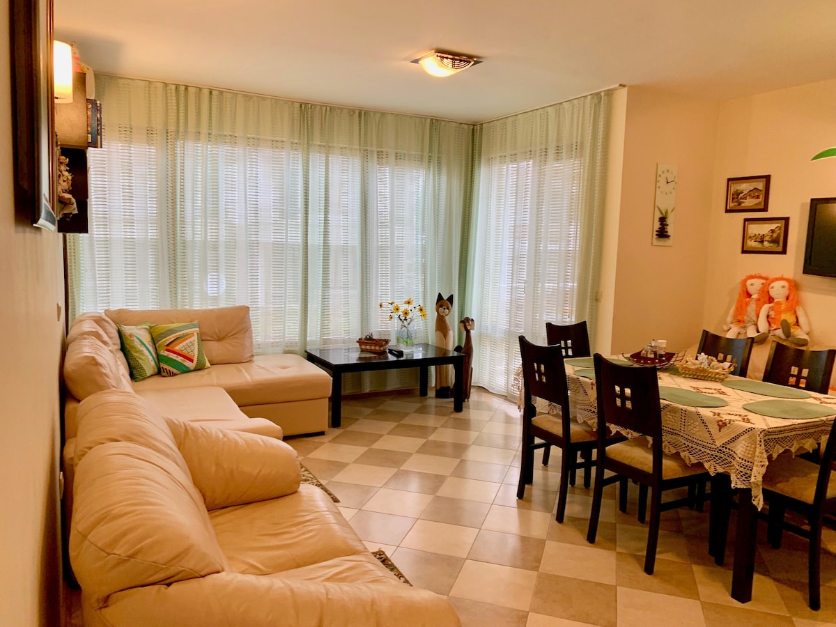 A bright living area features large windows adorned with sheer curtains, allowing natural light to fill the space. A comfortable sectional sofa is positioned near a coffee table, while a dining table with several chairs is visible, creating a welcoming environment for gatherings.