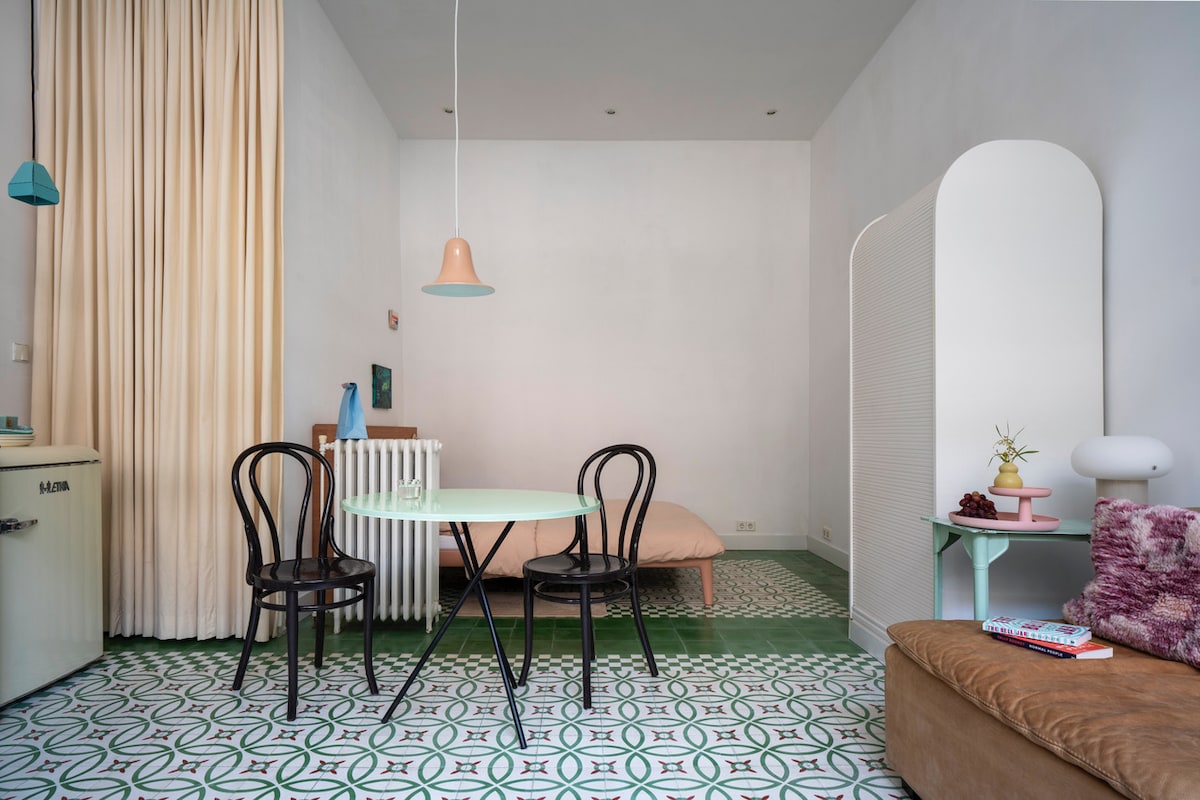 A cozy studio space features a light green table surrounded by two black chairs. A soft blush-colored bed is positioned against a wall, with a modern light fixture suspended above the table. The floor showcases a decorative tile pattern, enhancing the room's inviting atmosphere.