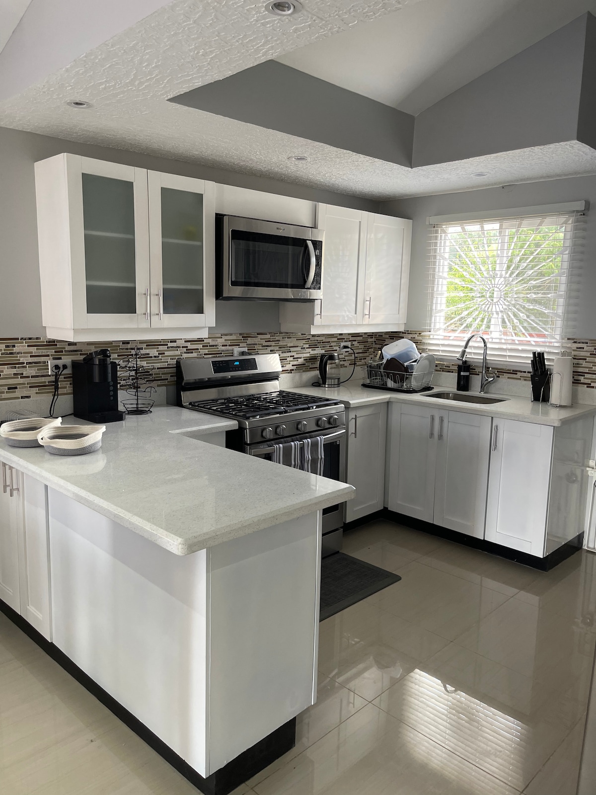 A modern kitchen features white cabinetry and stainless steel appliances. A gas stove is positioned alongside a microwave and refrigerator. Counter space is created with a light-colored granite surface. Natural light enters through a window adorned with decorative glass, enhancing the clean and functional design.