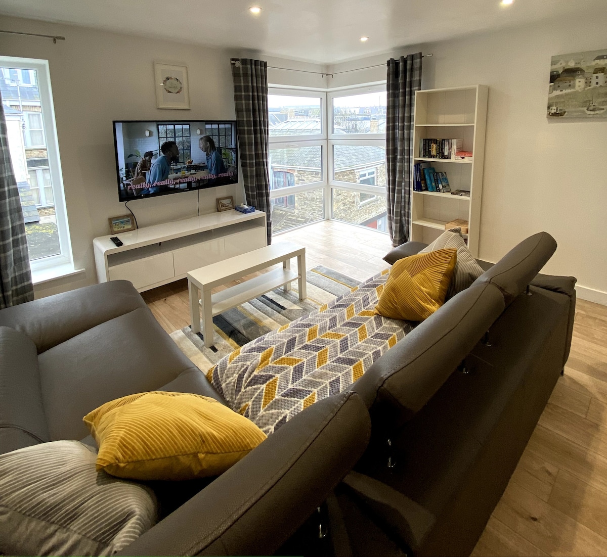 A spacious living area is highlighted by a large corner sofa adorned with yellow cushions. A white coffee table sits in front of the sofa, and a flat-screen TV mounted on the wall displays a scene from a show. Natural light is provided by large windows, partially covered by plaid curtains.