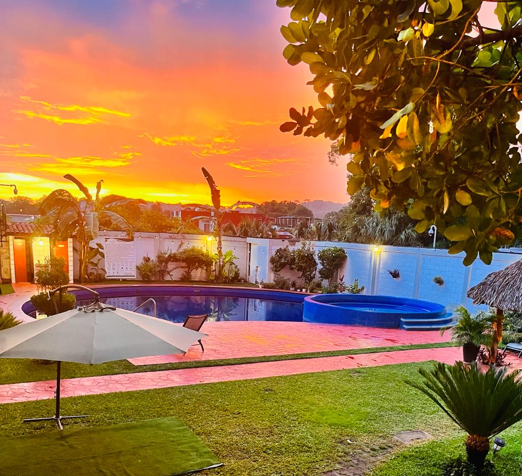 A tranquil outdoor space is showcased, featuring a spacious pool and a jacuzzi. Lush greenery surrounds the area, complemented by a sunset sky in hues of orange and pink. A shaded umbrella provides a resting spot, and a well-maintained lawn enhances the inviting atmosphere.