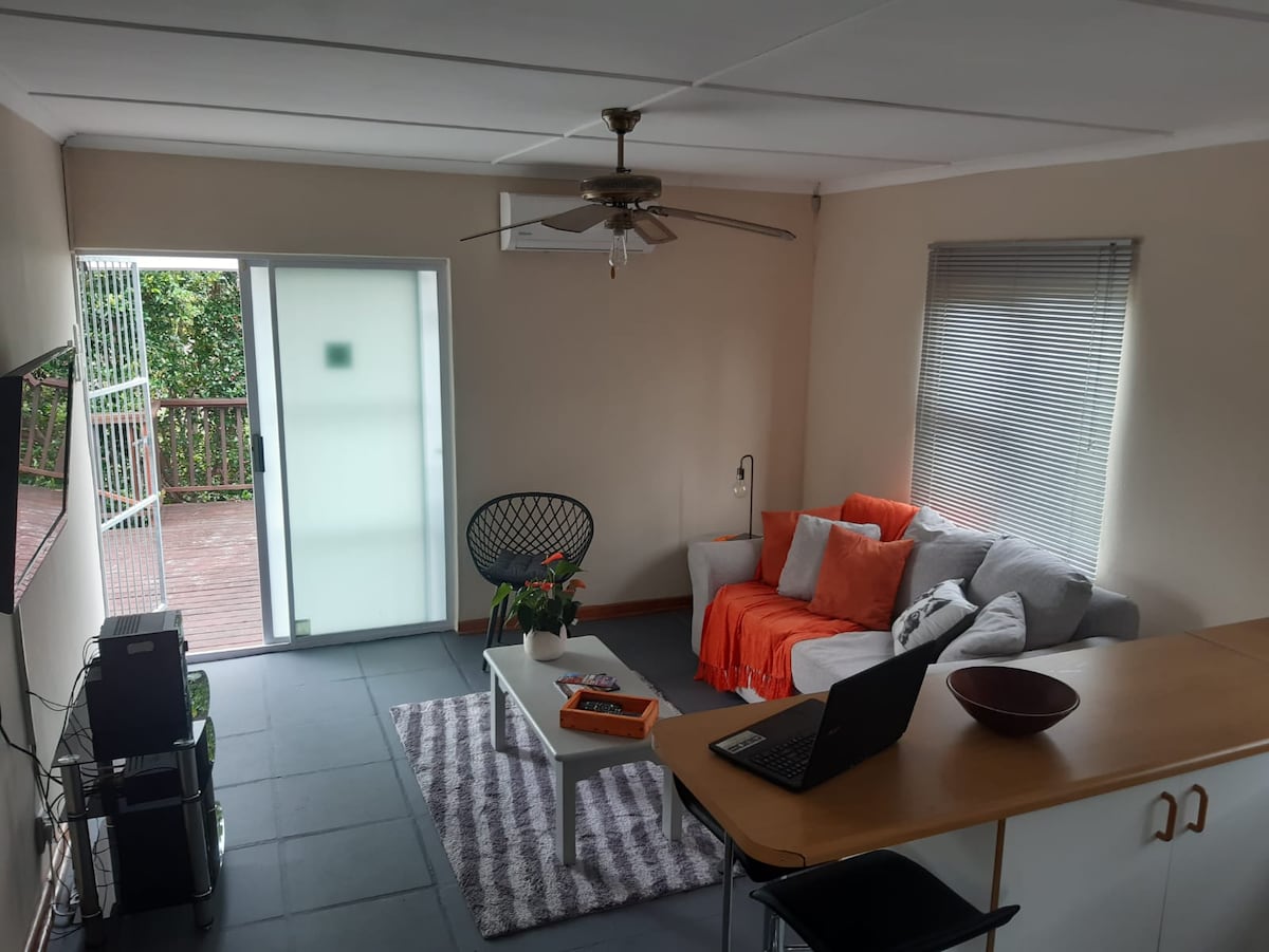 An inviting living space features a comfortable sofa adorned with orange throw pillows, complemented by a stylish chair. A coffee table sits in the center, while a laptop and a decorative bowl are placed on the nearby countertop. Natural light enters through the glass door leading to a small outdoor area.