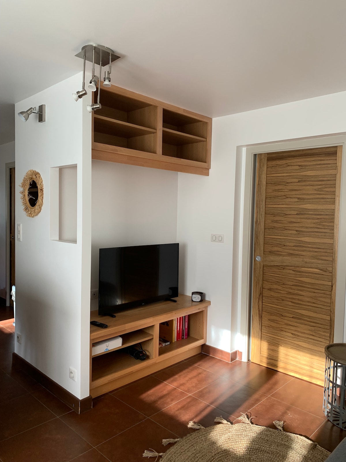 A media unit with wooden shelving displays a television and various items. The wall is bright and features an opening with a circular decorative mirror. A wooden door, with a textured finish, leads to another space, creating a sense of warmth.