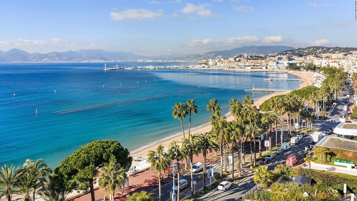 A panoramic view captures the coastline of Cannes, where turquoise waters gently lap against a sandy beach lined with palm trees. The vibrant promenade is visible, along with distant hills under a clear blue sky.