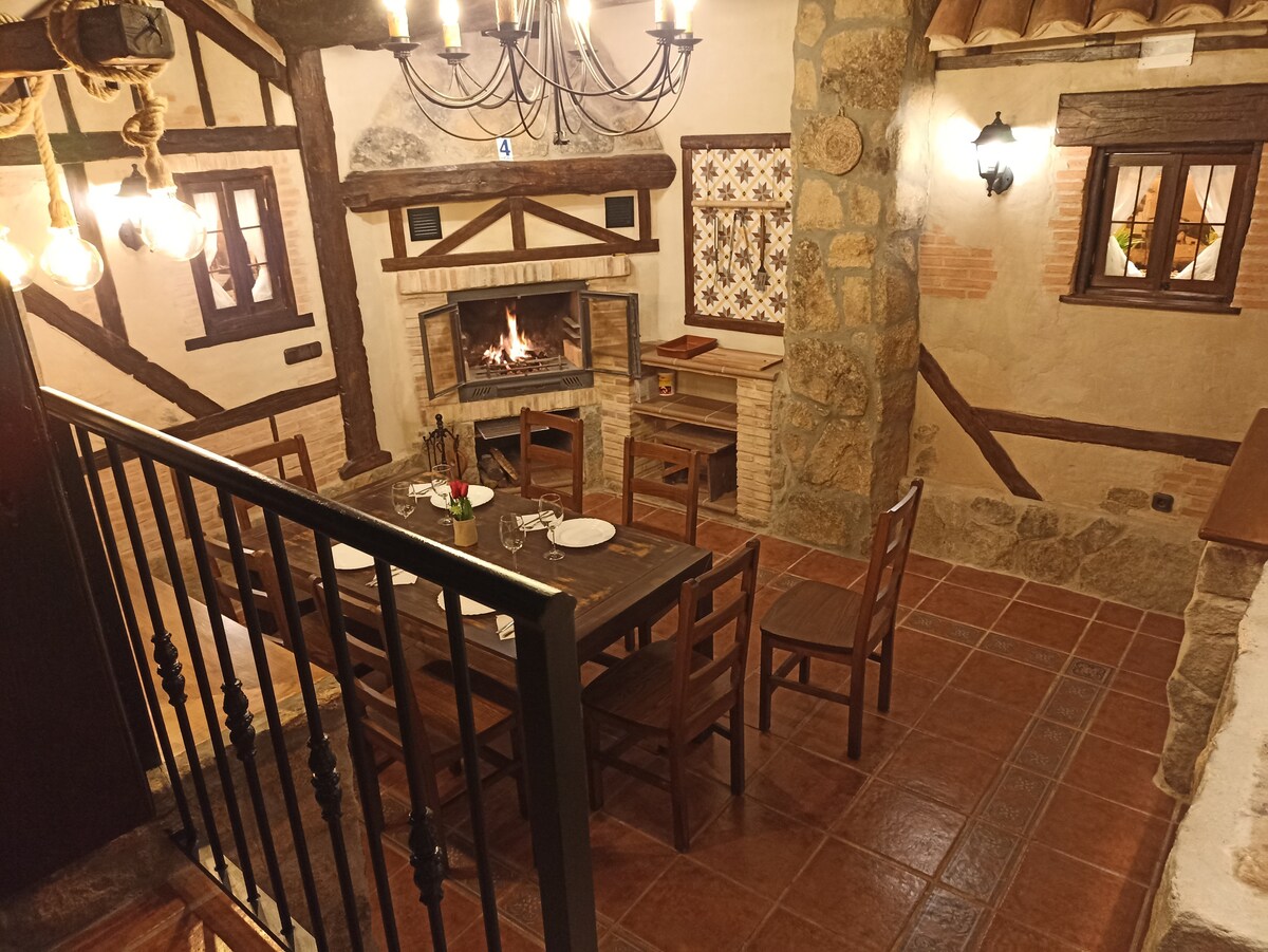 A rustic dining area features a wooden table surrounded by chairs, set against a stone fireplace that adds warmth. Soft lighting from overhead fixtures and wall sconces illuminates the space, highlighting the textured walls and charming architecture.