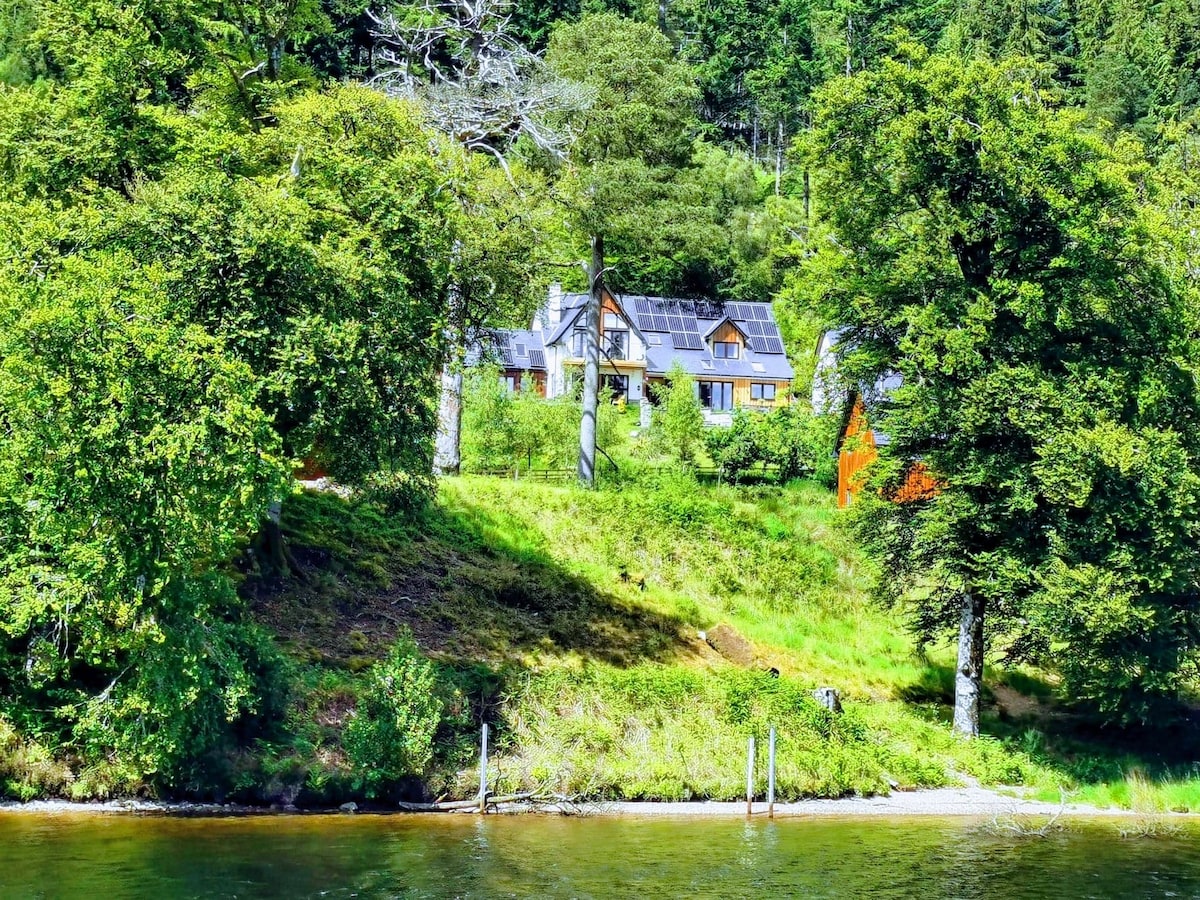 A family home is situated among lush greenery, with large windows and solar panels visible on the roof. Surrounding trees and grass create a natural setting, while the calm water reflects the vibrant colors of the landscape.