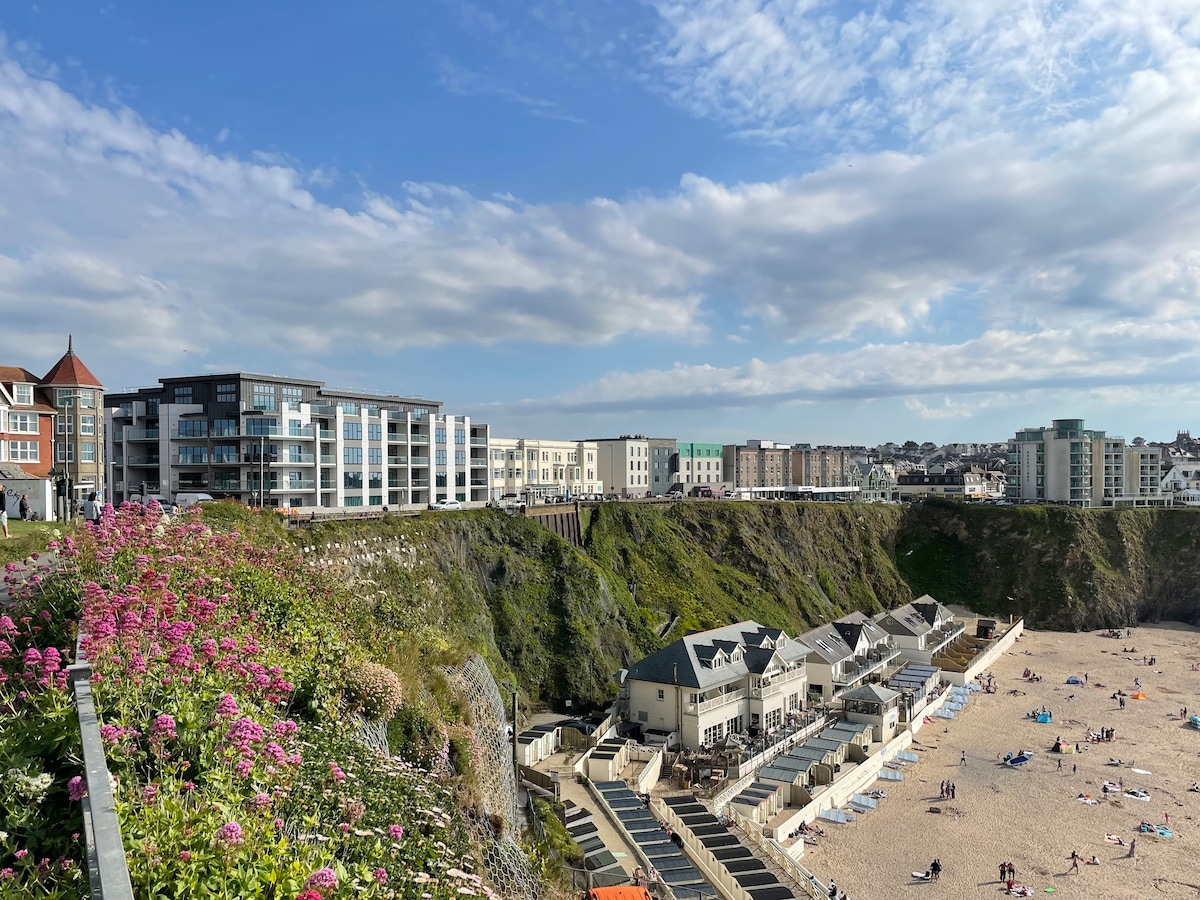 A panoramic view captures the coastline of Tolcarne Beach, featuring sandy areas with sunbathers and umbrellas. Colorful flowers in the foreground contrast with modern buildings and coastal cliffs in the background, illustrating the vibrant atmosphere of the Narrowcliff area.