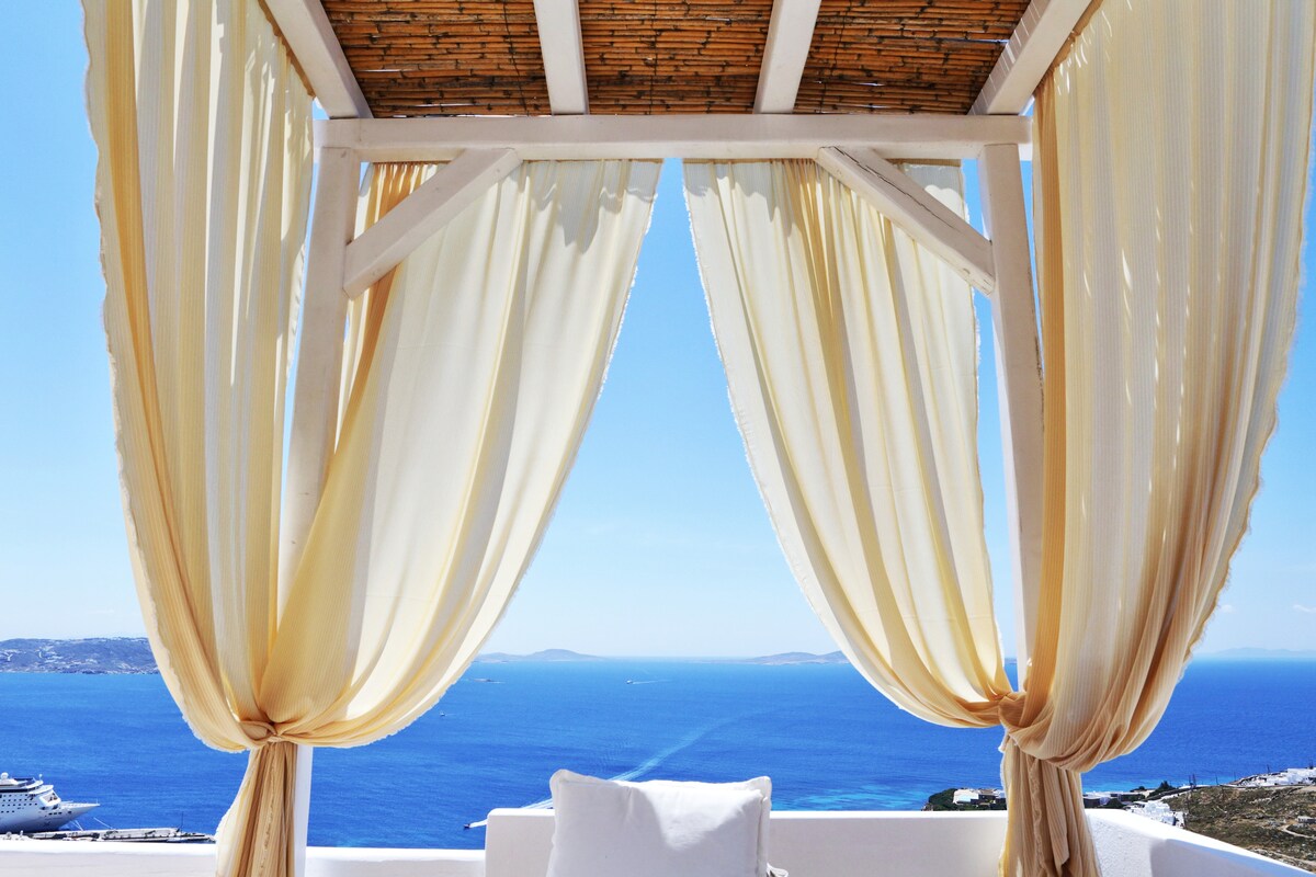 A white canopy is adorned with flowing sheer drapes, framing a panoramic view of the Aegean Sea. The bright blue water extends toward the horizon, with distant islands visible. A comfortable cushioned seating area is positioned below, inviting relaxation.