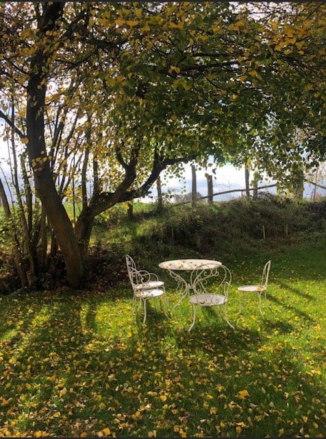 A small outdoor dining area is situated under a tree, featuring a round white table and two matching chairs. The ground is covered with yellow and green leaves, providing a natural setting. A rustic fence is visible in the background, adding to the peaceful atmosphere.