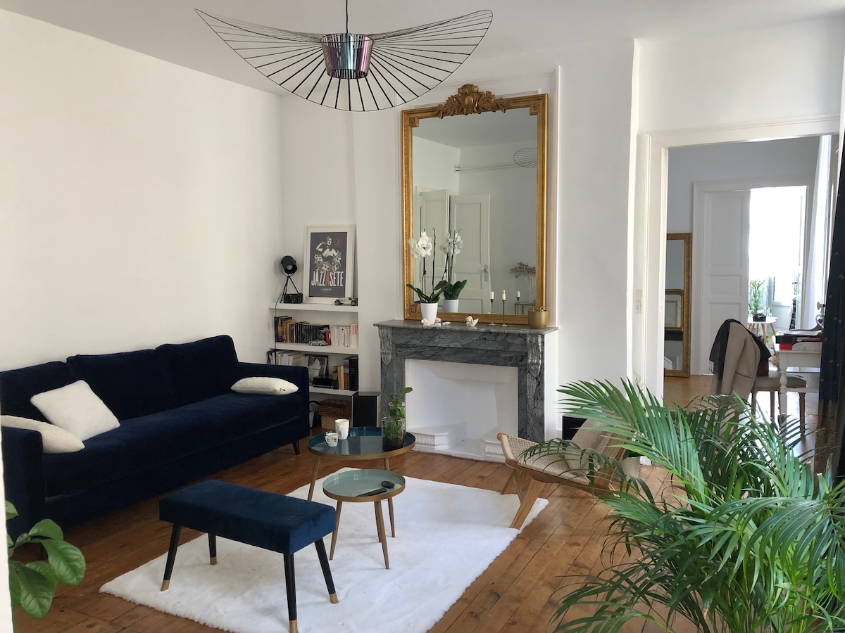 A neutral-toned living room features a deep blue sofa, a sleek coffee table, and a large area rug. A gold-framed mirror hangs above a marble fireplace, complemented by potted plants and artwork. Natural light comes through the adjacent room, enhancing the open feel.