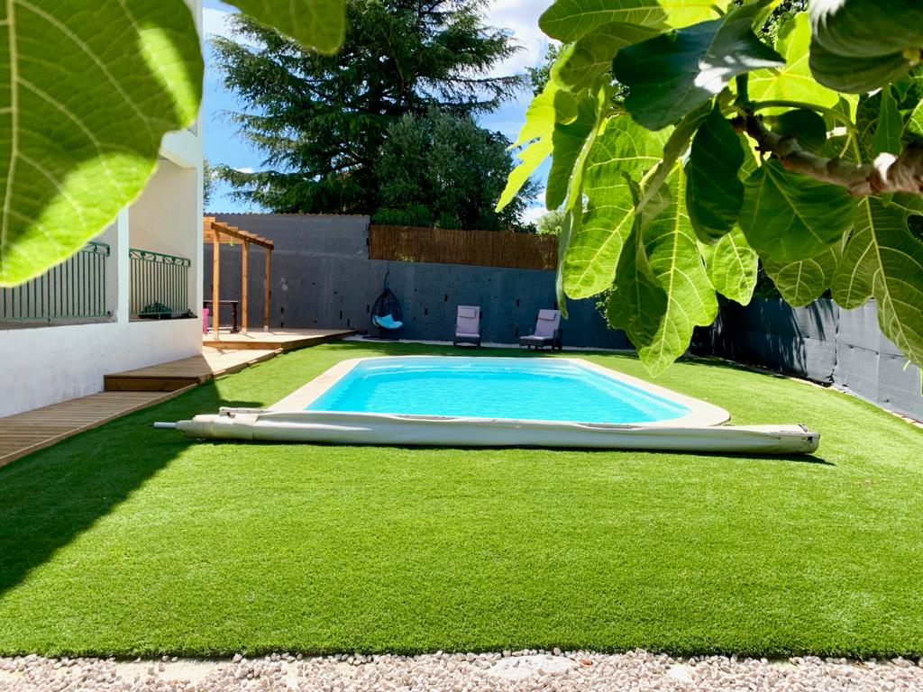 A serene outdoor space features a pool surrounded by well-maintained artificial grass and shade from leafy trees. Two loungers provide comfortable seating options, while a covered area suggests opportunities for relaxation and socializing. The tranquil environment is enhanced by the surrounding fencing.