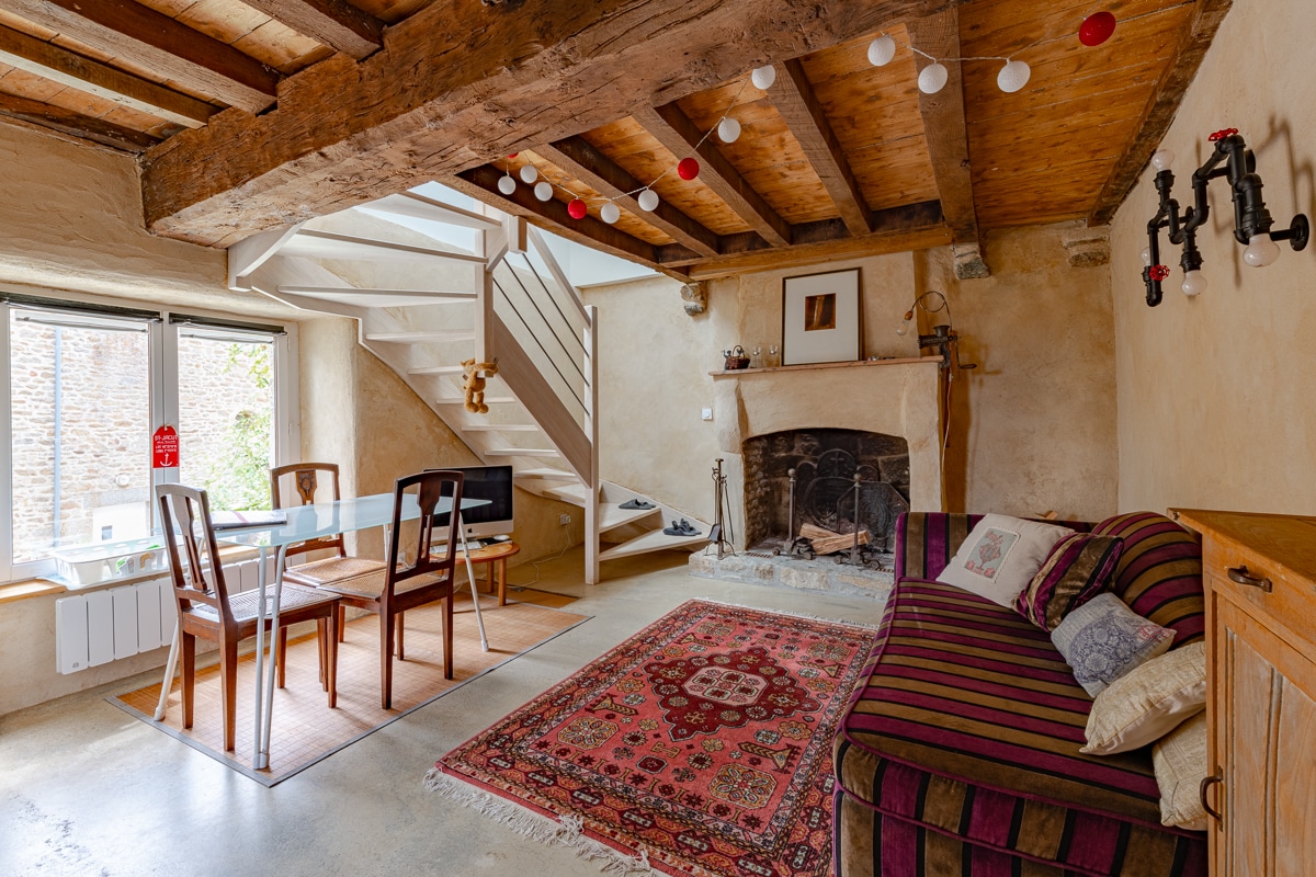 The living area features exposed wooden beams and a cozy ambiance. A striped sofa is positioned by the fireplace, complemented by a decorated rug on the floor. A dining table with four chairs is set near the window, allowing natural light to fill the space.