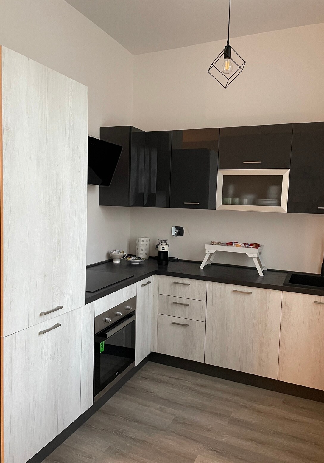 A modern kitchen is presented with sleek cabinetry in contrasting light and dark tones. Kitchen appliances, including an oven and coffee machine, are arranged efficiently. A subtle, geometric pendant light adds an elegant touch, while ample countertop space is provided for meal preparation.