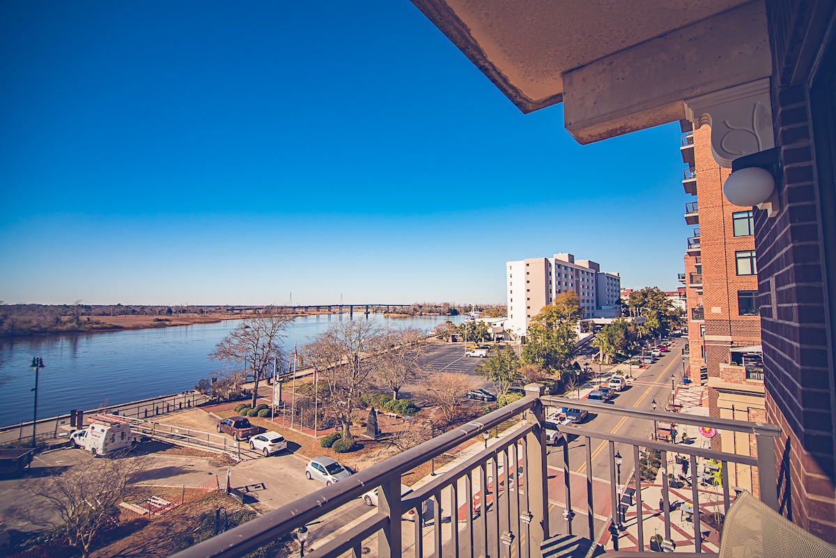 A balcony view overlooks the Cape Fear River, showcasing the water under a clear blue sky. The vibrant street below features trees and parked vehicles, while buildings line the horizon, creating an inviting urban atmosphere.