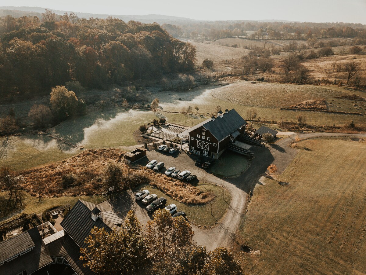 An aerial view captures a rural landscape featuring a large barn with a dark exterior, surrounded by green fields and patches of water. Vehicles are parked near the entrance, while trees line the horizon, enhancing the sense of tranquility in the countryside.