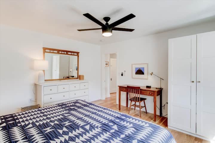 Primary bedroom view: large dresser with mirror, clothing wardrobe with hangers and extra bedding, writer's desk and charging station.