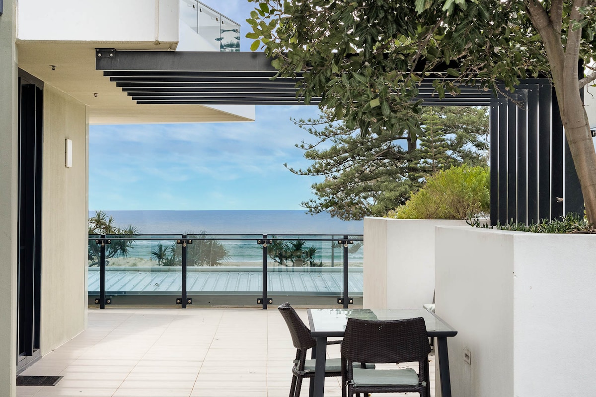 A private balcony is showcased, featuring a dining table set for outdoor meals. Lush greenery frames the space, while glimpses of the ocean are visible in the background, enhancing the serene coastal environment.