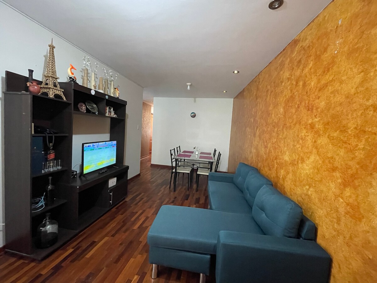 A modern living area features a dark blue sectional sofa adjacent to a wall-mounted television. A stylish black shelf displays decorative items, while a dining set for four is positioned in the background against a textured orange wall. Natural light illuminates the wood flooring.