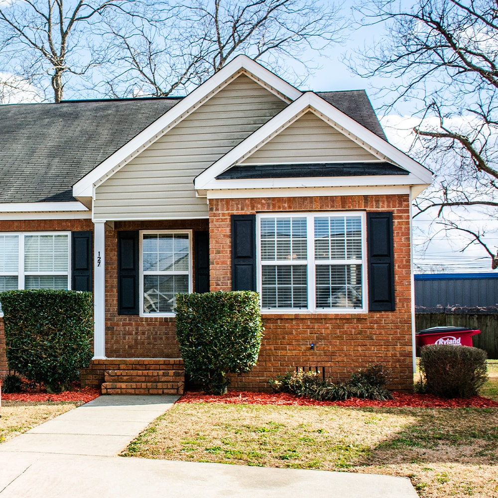 The exterior of the condo features a brick facade with light-colored siding and black shutters. A pathway leads to the entrance, bordered by neatly trimmed shrubs. The window allows natural light into the living space, complemented by a gravel driveway offering ample parking.