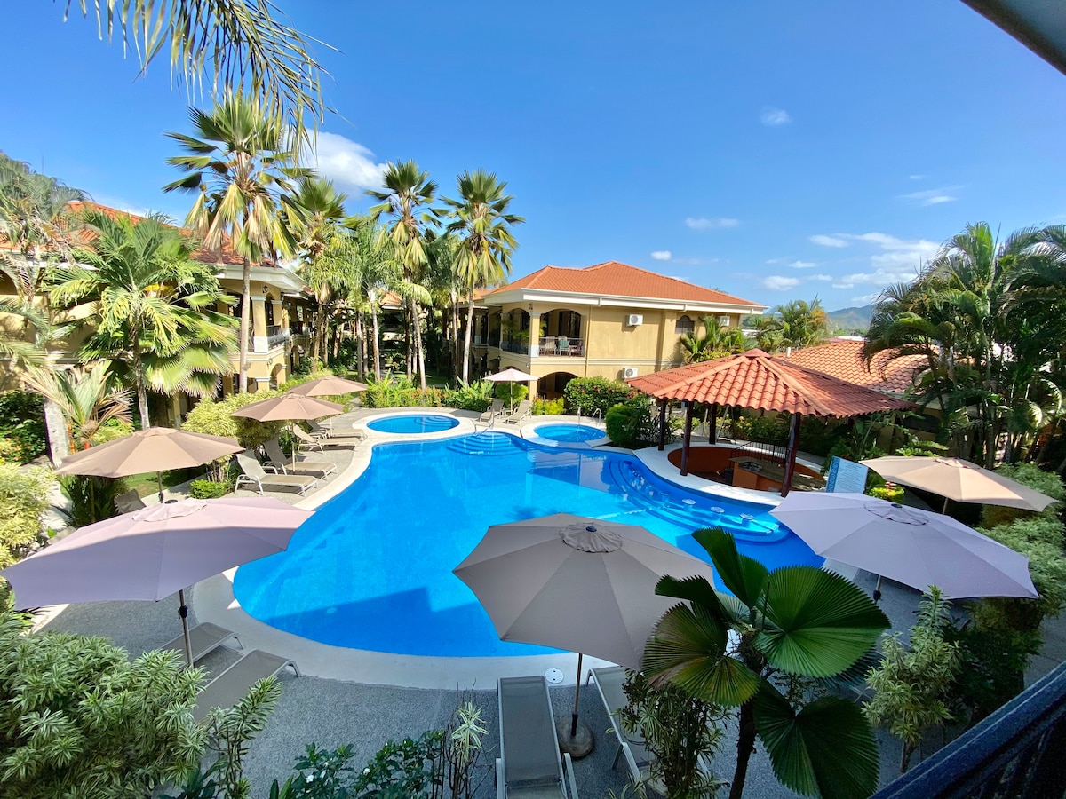 A inviting outdoor pool area is surrounded by lush palm trees and tropical plants. Sun loungers are placed beside the pool, with large umbrellas providing shade. A covered seating area is visible nearby, offering a space for relaxation under a tiled roof.