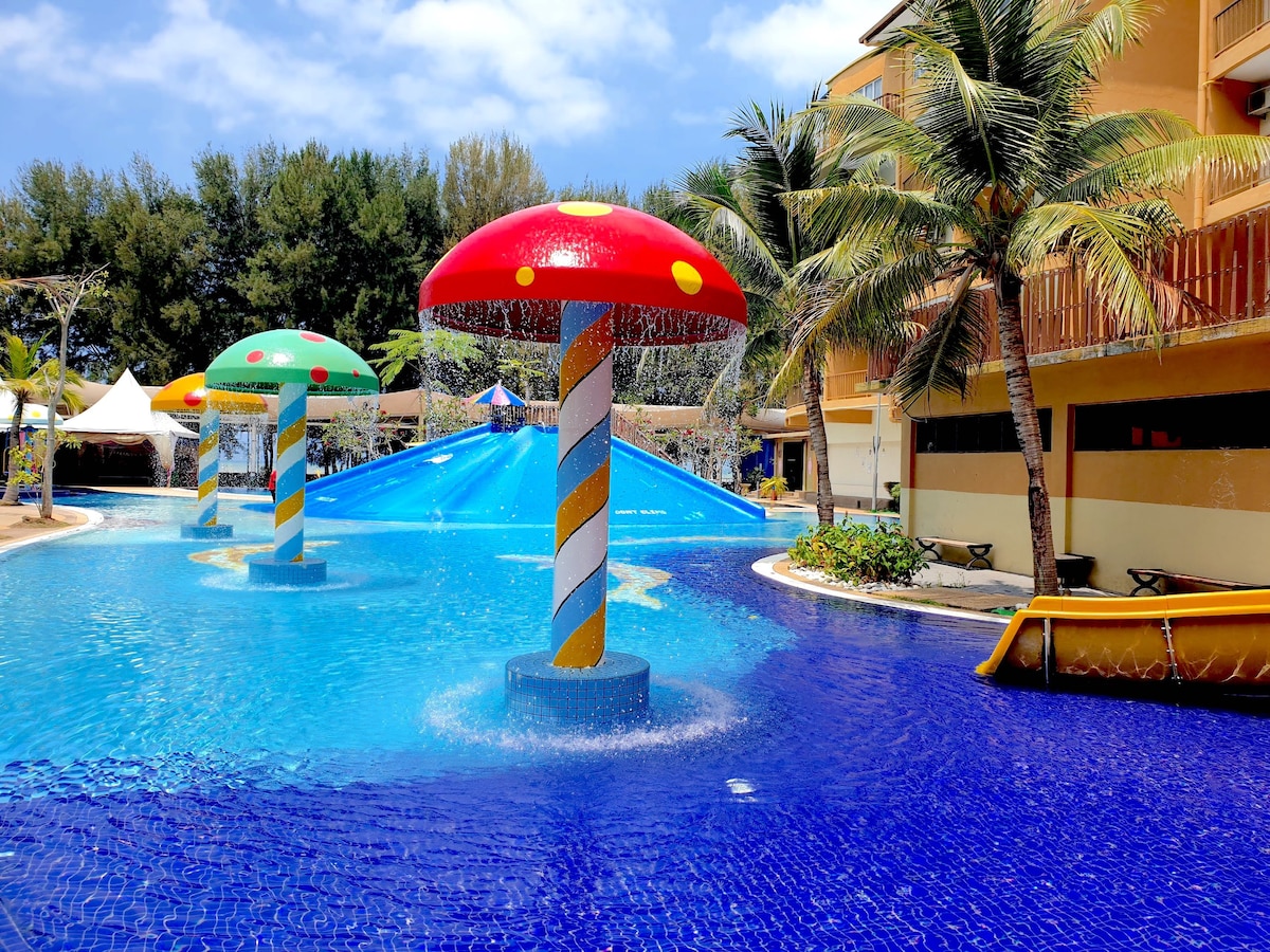 A vibrant water theme park area features colorful mushroom-shaped fountains amidst a large blue pool. Lush palm trees are visible along the edges, and bright blue skies complement the cheerful atmosphere created by the playful structures and landscaping.
