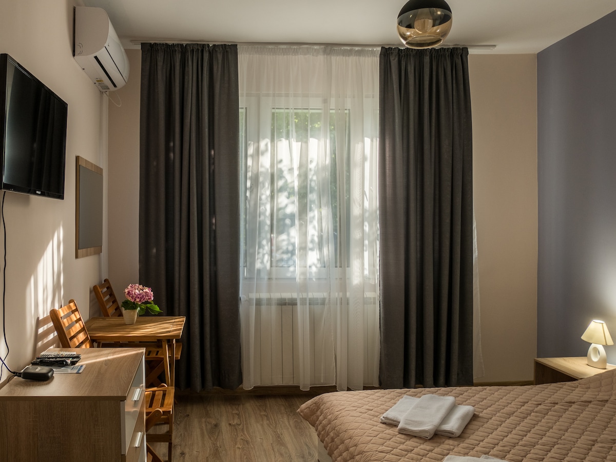 A spacious guest room is shown, featuring light walls and wooden flooring. A bed with neutral bedding is positioned in the foreground, while a small table and chairs are placed by the window, partially covered by sheer curtains. A flat-screen TV is mounted on the wall.