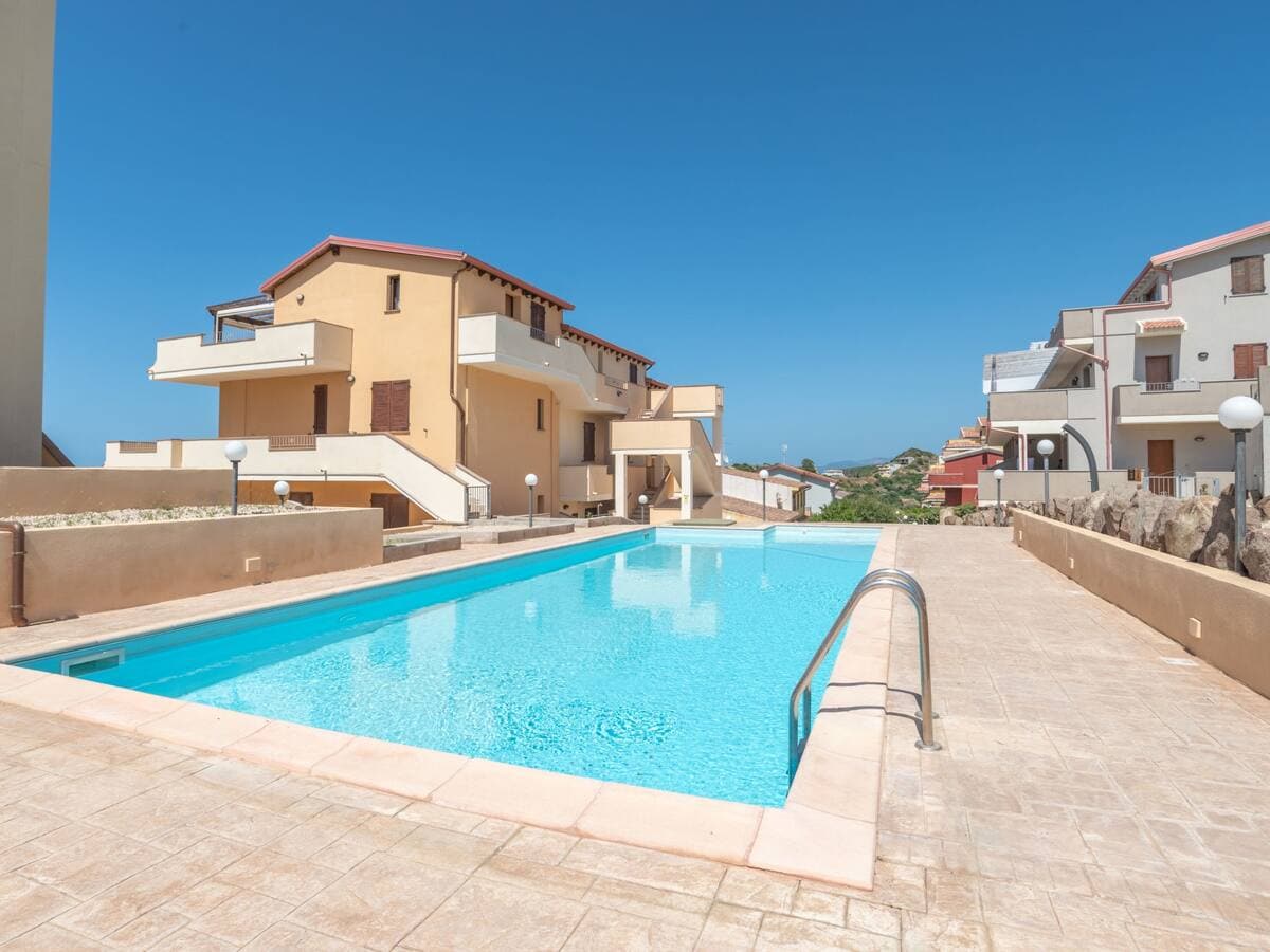 A shared swimming pool is presented, featuring clear blue water under a bright sky. The surrounding area is paved and includes a well-defined border. Nearby, buildings with neutral-colored exteriors are visible, creating a relaxed atmosphere for guests seeking leisure.