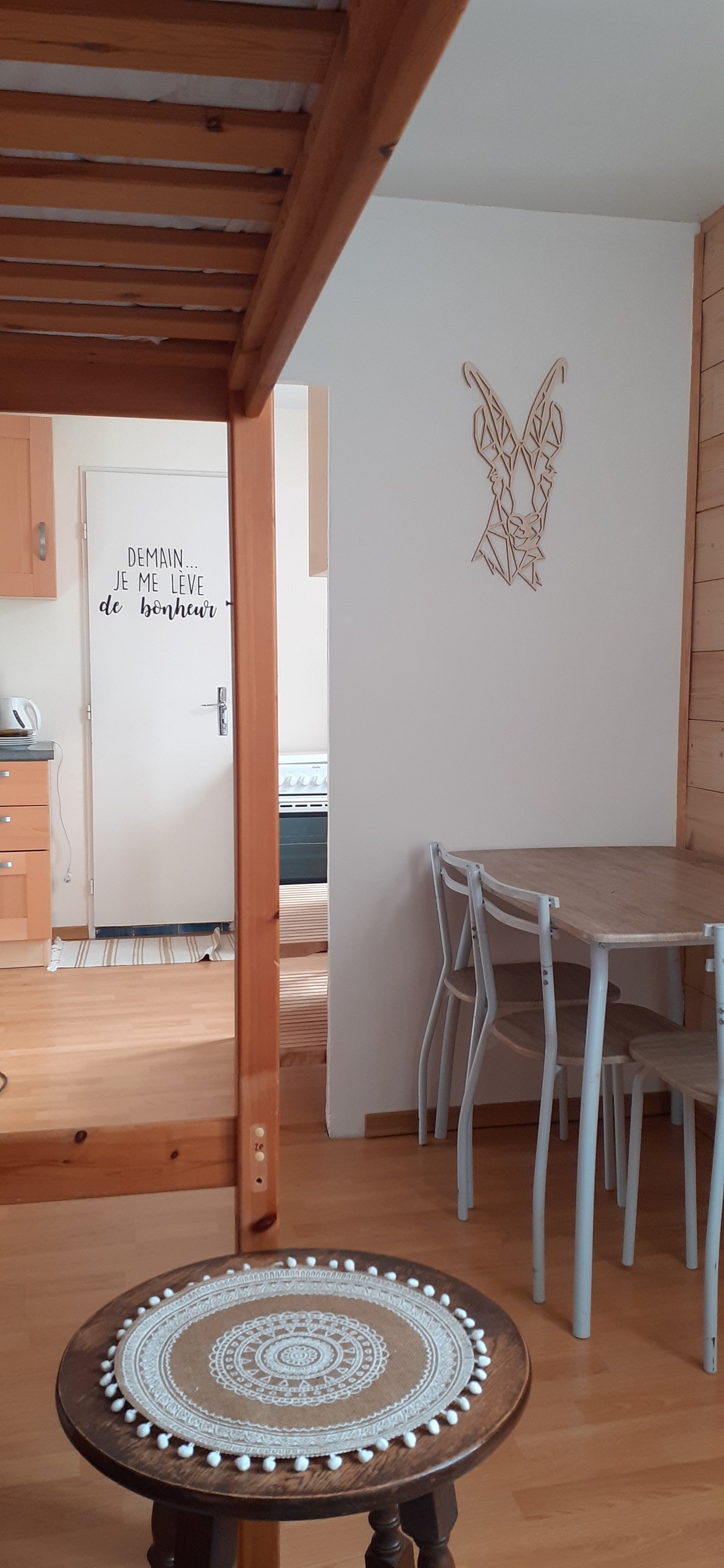 A wooden staircase leads to a cozy living area featuring a small dining table with four chairs. A decorative wall piece adds character to one side, while the kitchen area is visible in the background. Light-colored wooden walls complement the warm atmosphere.