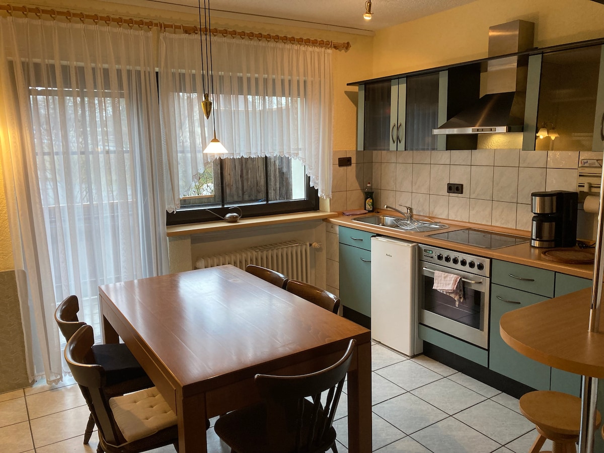 A well-equipped kitchen area features a wooden dining table with several chairs, complemented by blue cabinetry. A window dressed with sheer curtains allows natural light to illuminate the space. Kitchen appliances, including an oven and coffee maker, are neatly arranged along the counter.
