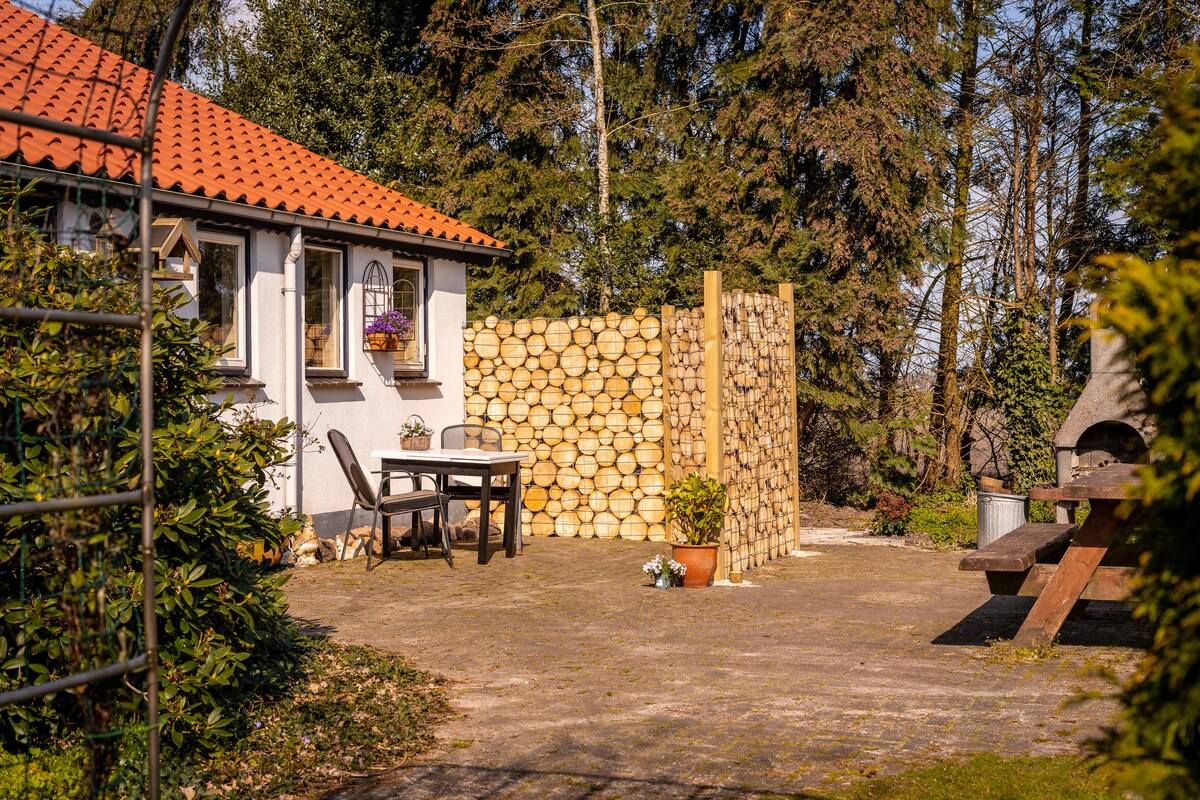A charming patio area features a wooden table and chairs, surrounded by greenery and bordered by a natural wooden privacy screen. The warm sunlight casts gentle shadows, inviting relaxation in a peaceful outdoor setting.