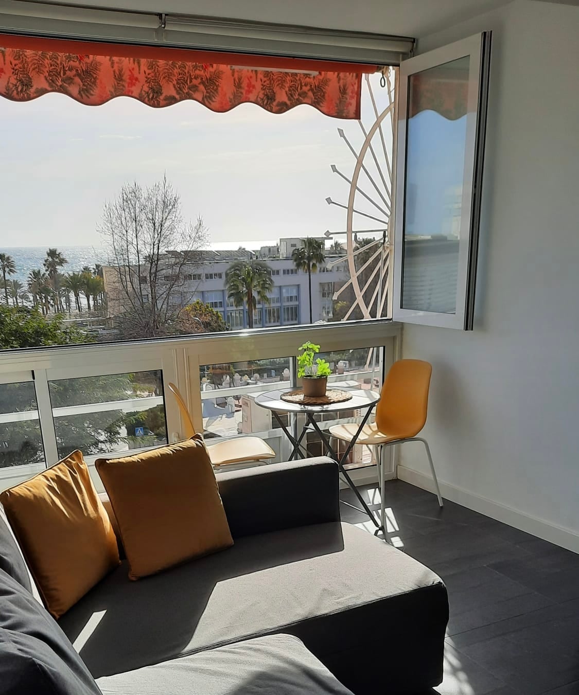 A sunlit seating area features a gray sofa adorned with orange cushions. A small round table holds a potted plant, and two yellow chairs are placed nearby. A large window offers views of palm trees and the ocean in the distance, complemented by a red awning.