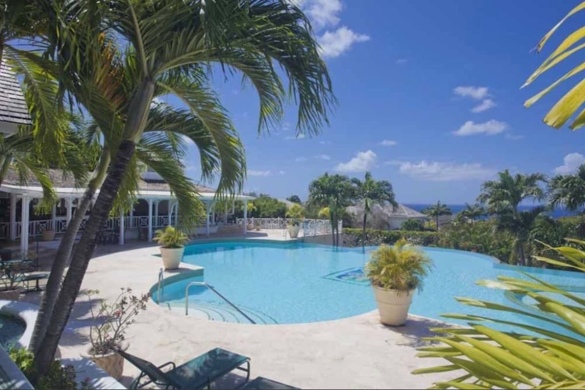 A large outdoor swimming pool with a serene blue interior is complemented by surrounding palm trees and lounge chairs. The pool area is framed by a patio that leads to a welcoming clubhouse and offers views of distant hills and a clear blue sky.