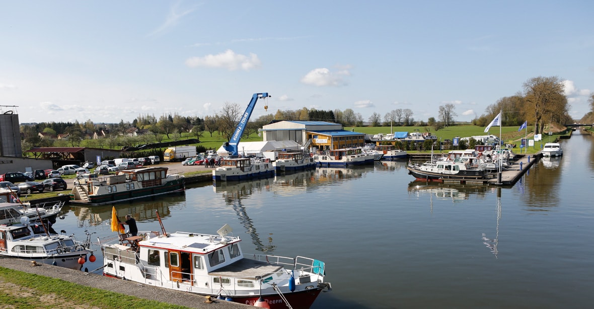 The image displays a serene waterfront scene featuring a variety of boats docked along a calm river. Lush greenery surrounds the area, with a number of buildings visible in the background. A crane is positioned on the shore, contributing to the marina's activity.