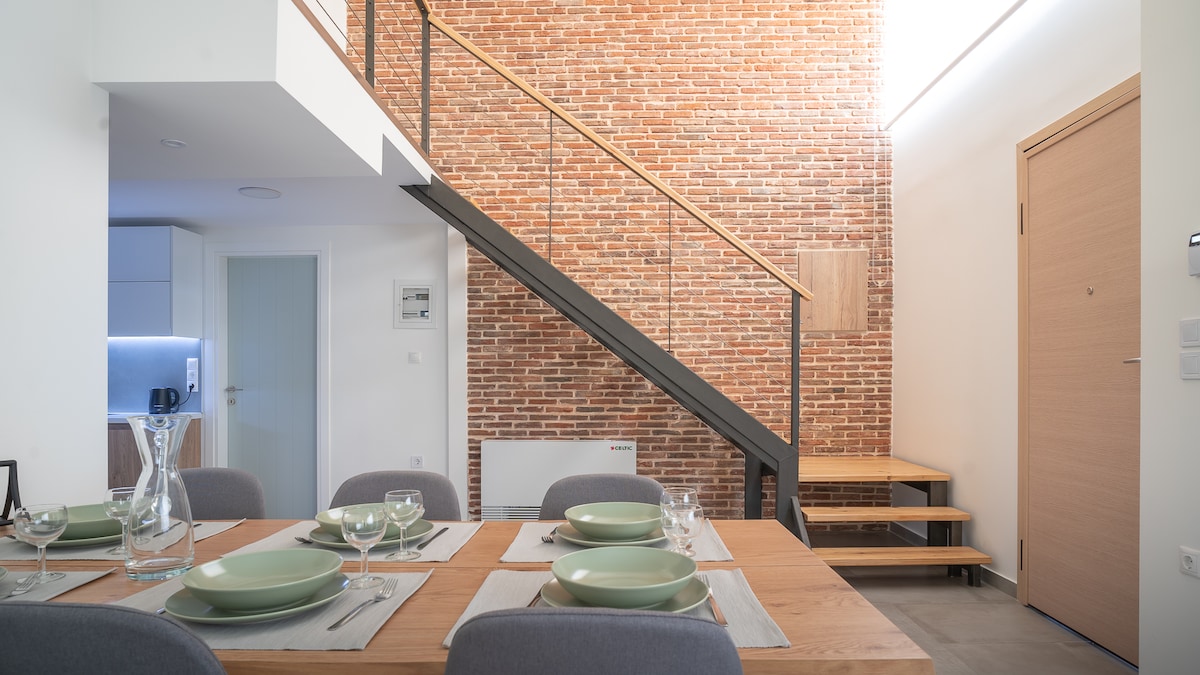 A modern dining area features a wooden table set with pale green plates and glassware, complemented by grey upholstered chairs. A staircase with a wooden handrail leads to the upper level, alongside a textured red brick wall that enhances the space's design.