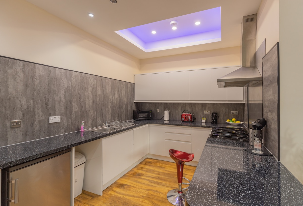 A modern kitchen is displayed with sleek gray cabinetry and a dark stone countertop. A red bar stool is positioned at the countertop, and essential appliances, including a microwave and kettle, are visible. Soft lighting enhances the open feel of the space.