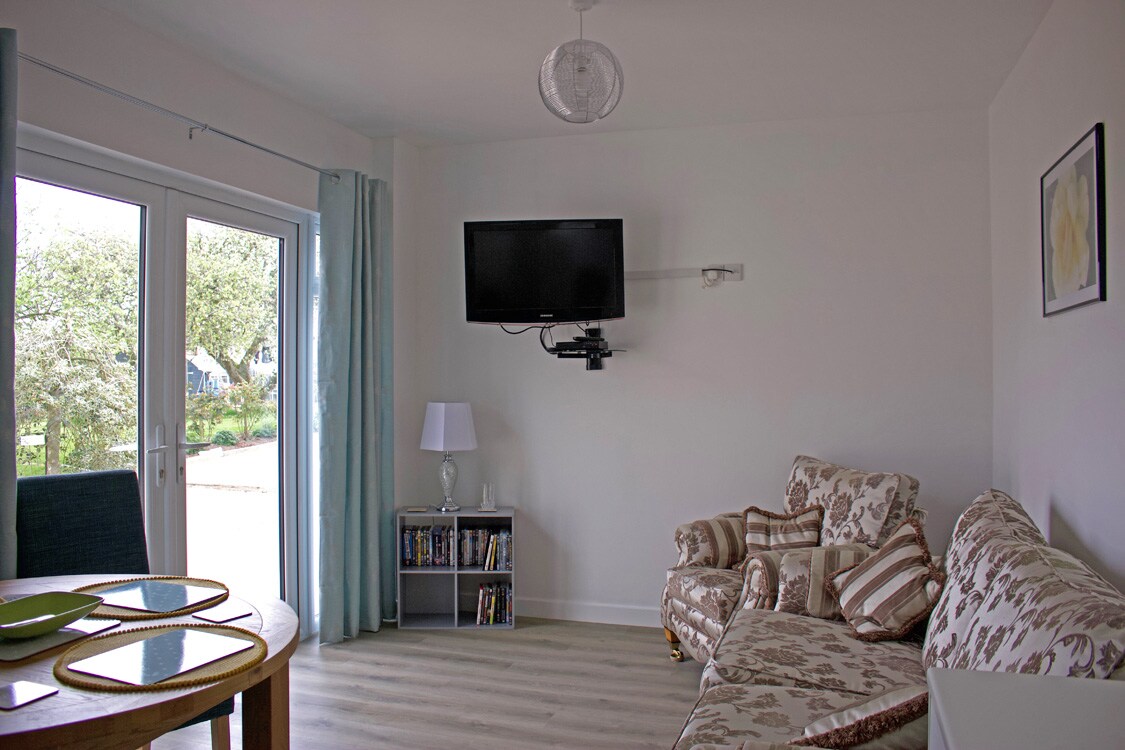 A light-filled living area is presented with soft grey walls and a wooden floor. A floral-patterned sofa offers comfortable seating, complemented by a round table set with plates. A wall-mounted TV is positioned above a small shelf containing DVDs, while large doors open to the garden.
