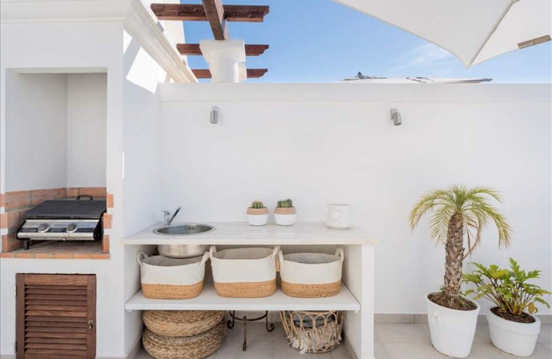 An outdoor kitchen area is presented with a built-in grill, a sink, and a countertop finished in light stone. Storage baskets are neatly arranged beneath the counter. Potted plants, including a small palm, add a touch of greenery against the white walls.