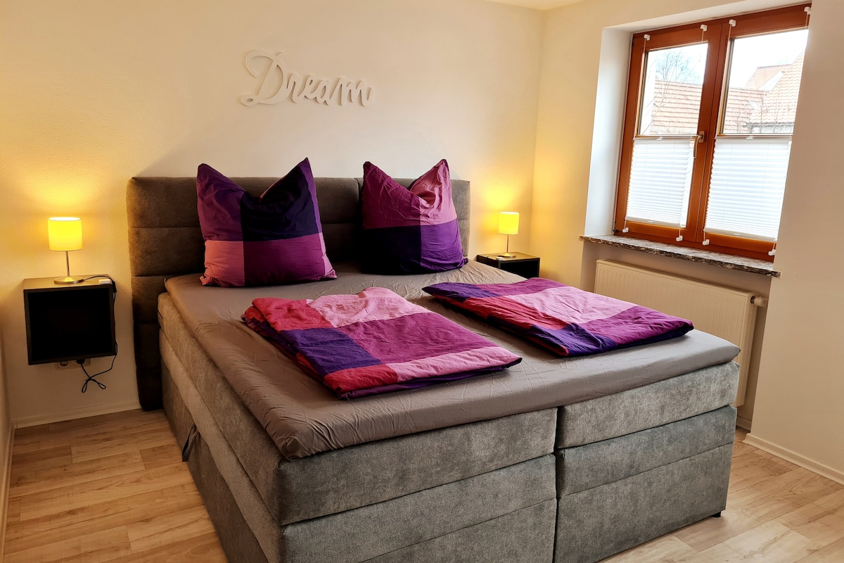 A spacious bed is adorned with colorful purple and pink bedding. Two bedside lamps provide soft lighting, and a decorative wall feature reads 'DREAM.' Natural light enters through a window dressed with white blinds, illuminating the serene room.