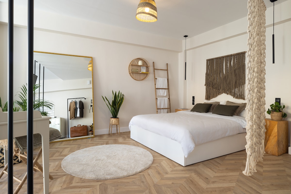 A bright bedroom features a spacious bed with neutral bedding and decorative pillows. A large round rug complements the wood flooring. A mirror and ladder shelf are positioned against the wall, alongside indoor plants that enhance the natural feel of the space.