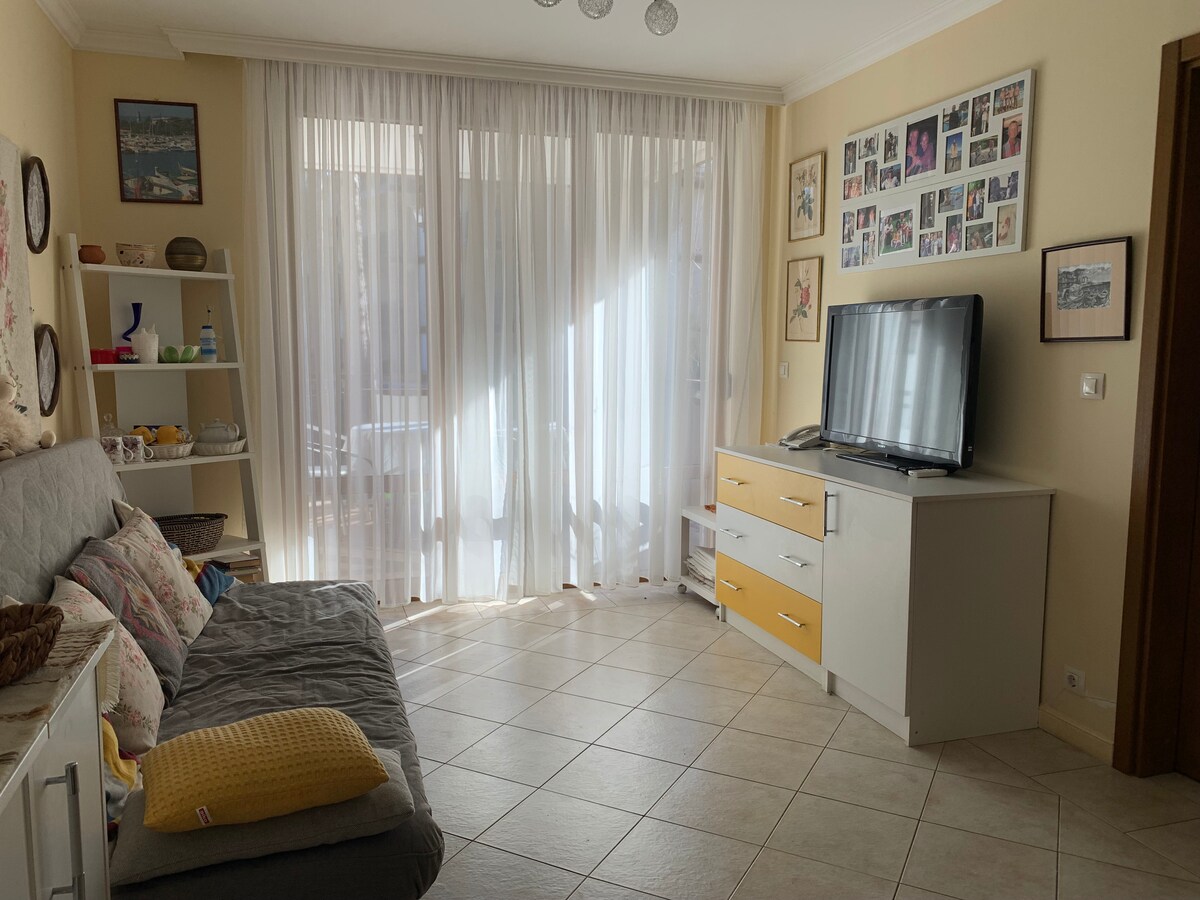 A bright living area features a light-colored sofa bed against a sunny window, adorned with sheer curtains that gently filter the light. A modern TV unit with storage sits opposite, while walls are decorated with framed photographs, adding a personal touch to the space.