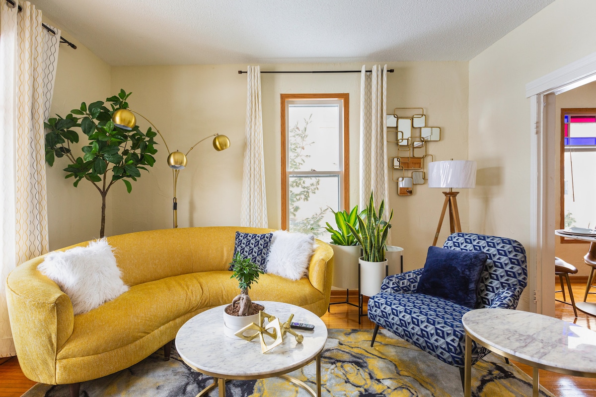 A bright living area features a curved yellow sofa complemented by white throw pillows. A patterned blue chair sits beside two round marble tables atop a colorful area rug. A large plant adds greenery, while natural light streams through a window with sheer curtains.
