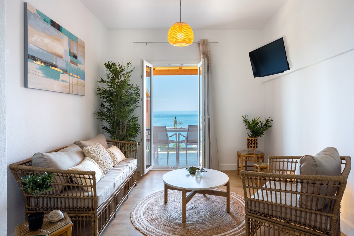 A comfortable living area features a light color scheme with two rattan sofas arranged around a circular coffee table. A natural fiber rug lies beneath, while large glass doors open to reveal a view of the sea. A flat-screen TV hangs on the wall.