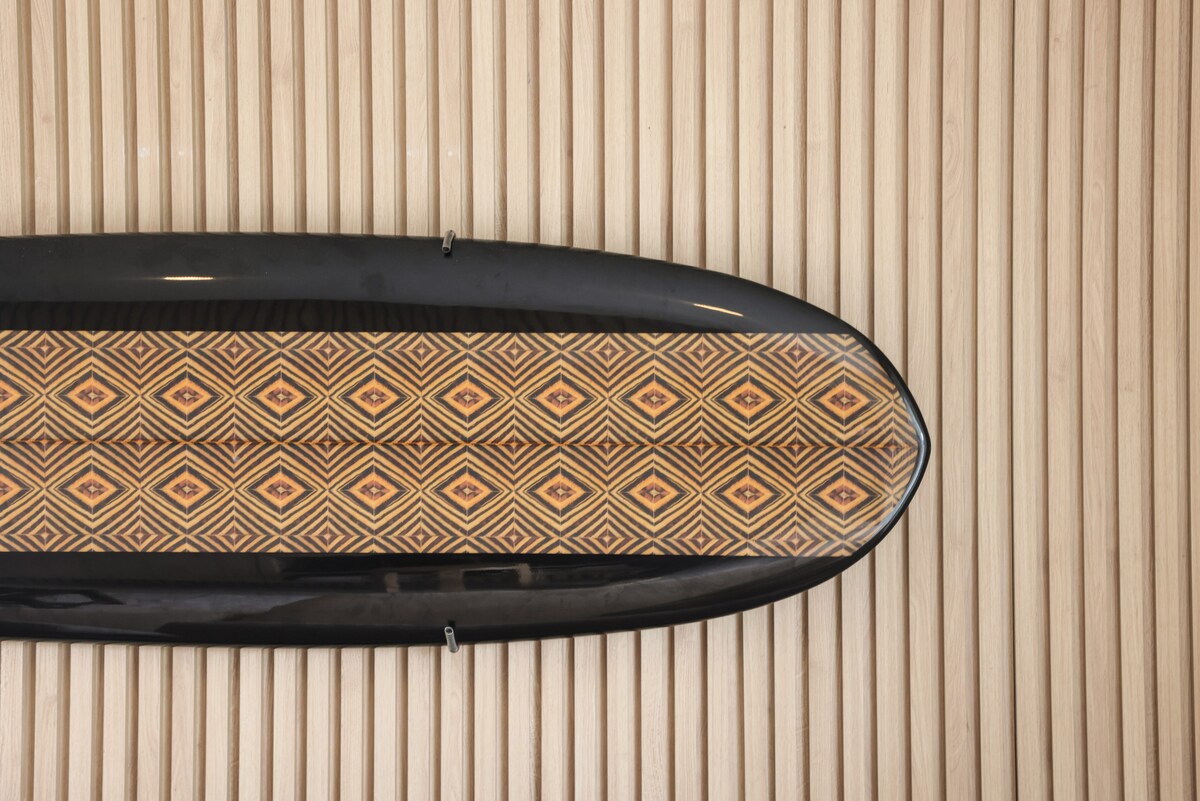 A surfboard with a geometric pattern in warm tones is displayed against a backdrop of textured wooden panels. The combination of the dark edges and the intricate design creates a striking visual element.