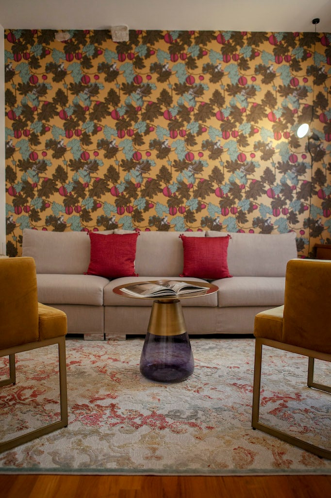 A living area features a beige sofa adorned with two pink cushions, complemented by two mustard yellow chairs. A glass tabletop sits on a decorative, floral rug, with a magazine displayed on it. The wall is covered in a vibrant, patterned wallpaper.