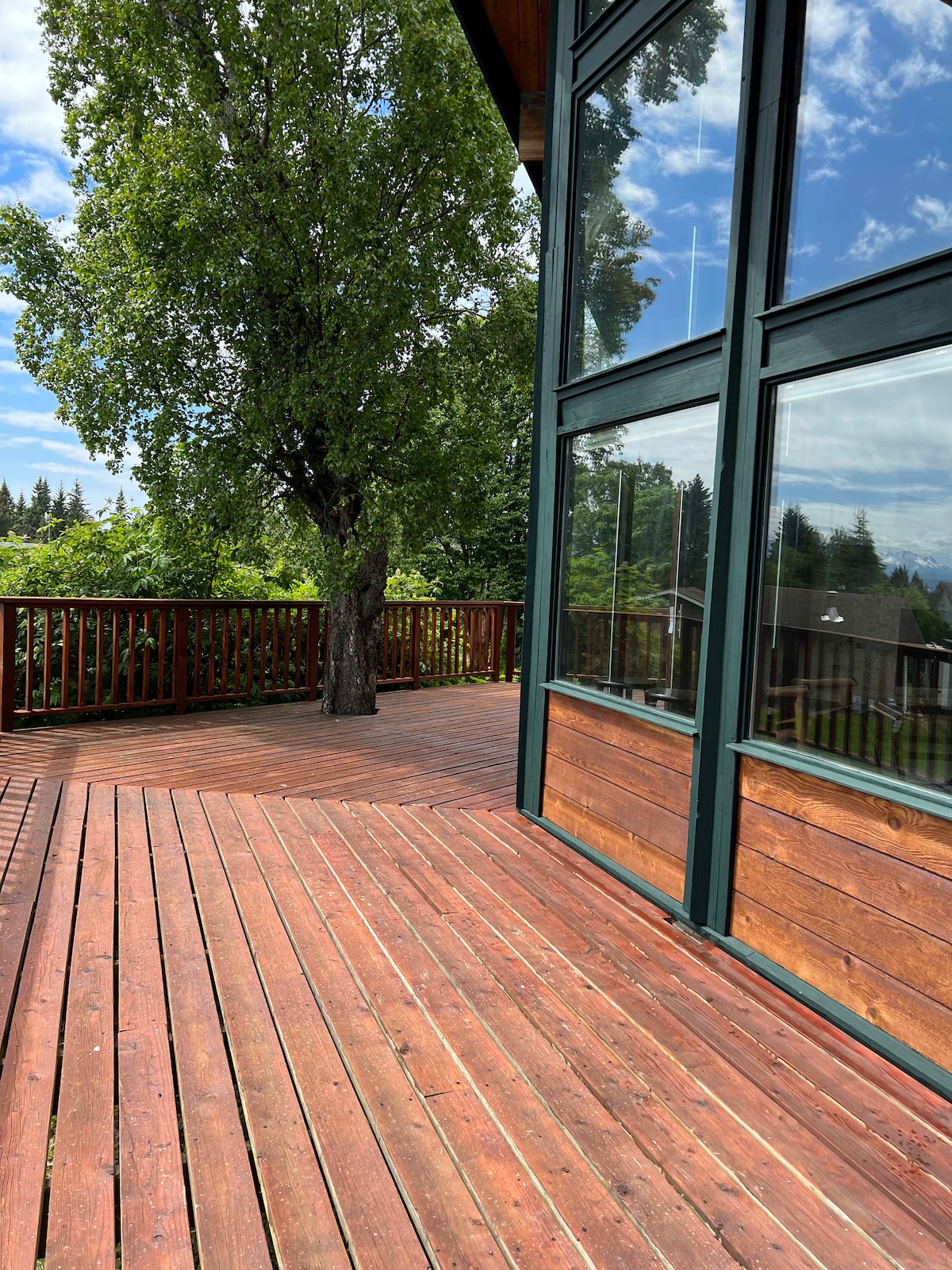 A spacious deck features rich wood flooring and large windows showcasing scenic views of the surrounding greenery. A single tree provides shade, contributing to the serene outdoor atmosphere. The deck wraps around the main level, offering ample space for relaxation.